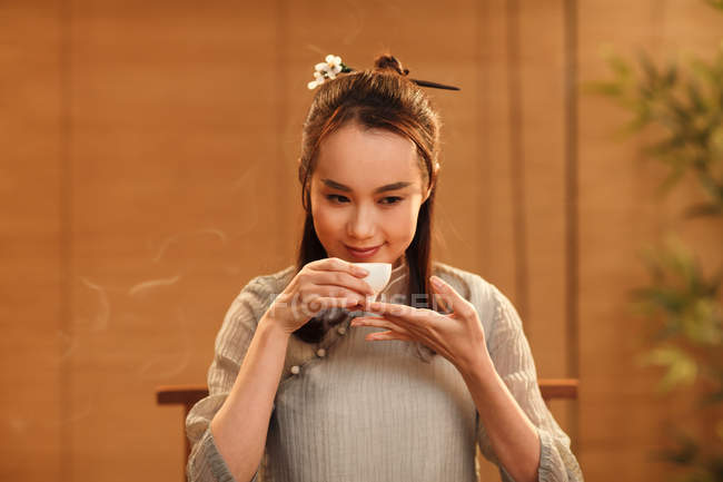 Hermosa sonriente joven mujer china sosteniendo taza blanca y oliendo té aromático - foto de stock