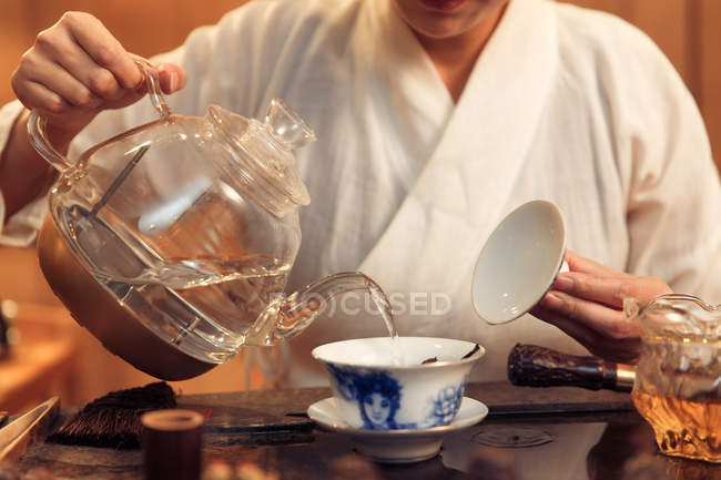 Tiro cortado de mulher segurando bule e derramando água em recipiente de porcelana — Fotografia de Stock
