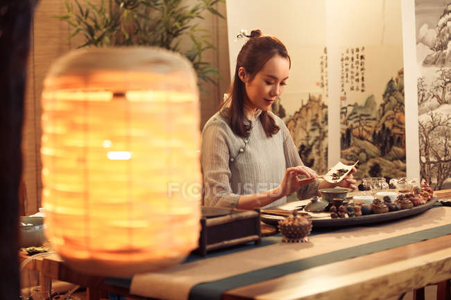 Крупный план освещённого фонаря на переднем плане и улыбающаяся молодая китаянка, заваривающая чай позади — стоковое фото