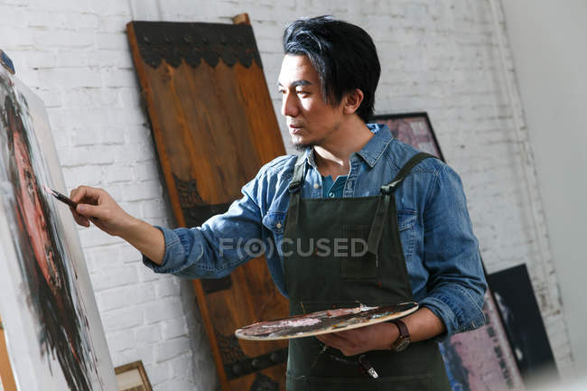 Bel homme asiatique dans tablier tenant palette et tableau de peinture dans le studio d'art — Photo de stock