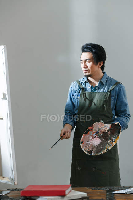 Concentrated young asian artist in apron holding palette and painting picture in studio — Stock Photo