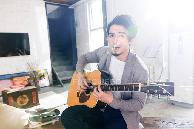 Schöner asiatischer Mann spielt Akustikgitarre und schaut zu Hause in die Kamera — Stockfoto