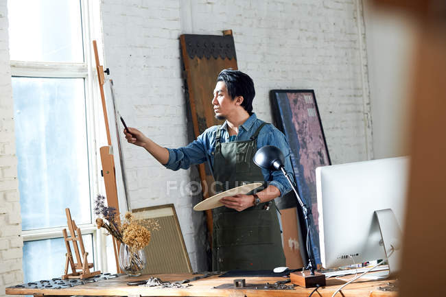 Сфокусированный художник держит палитру и картину в студии — стоковое фото