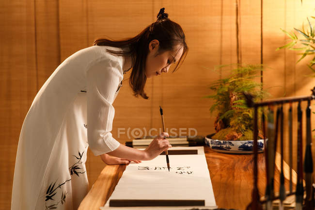 Vista lateral de bela jovem asiática mulher segurando escova e escrevendo caracteres chineses — Fotografia de Stock