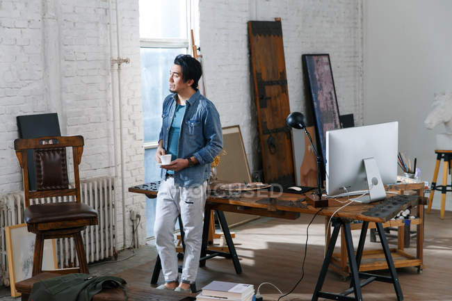 Pensive asian man holding cup of coffee and looking away in art studio — Stock Photo