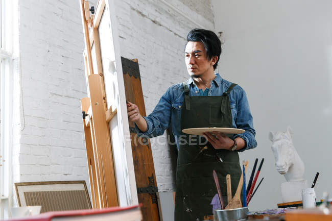 Concentrated young asian artist in apron holding palette and painting picture in studio — Stock Photo