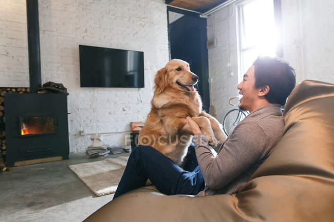 Seitenansicht eines glücklichen asiatischen Mannes, der auf einem Sitzsack-Stuhl sitzt und zu Hause mit Hund spielt — Stockfoto
