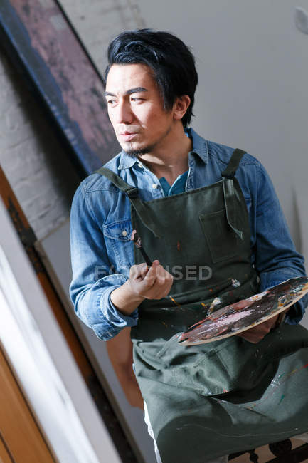 Serious male artist in apron holding palette and looking at picture in studio — Stock Photo