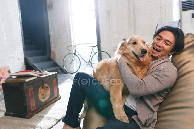 Glückliche asiatische Mann sitzt auf Bohnensack Stuhl und spielt mit Hund zu Hause — Stockfoto