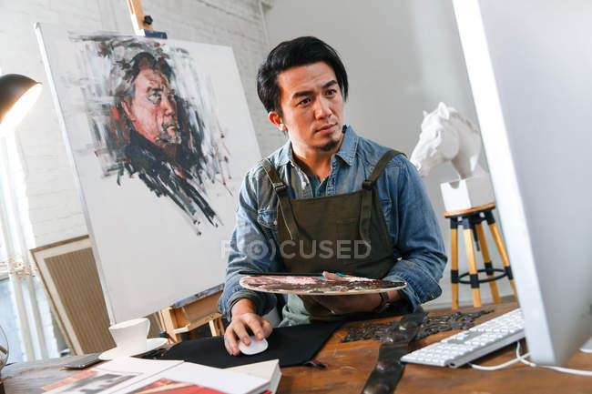 Bello asiatico pittore holding tavolozza e utilizzando desktop computer in studio — Foto stock