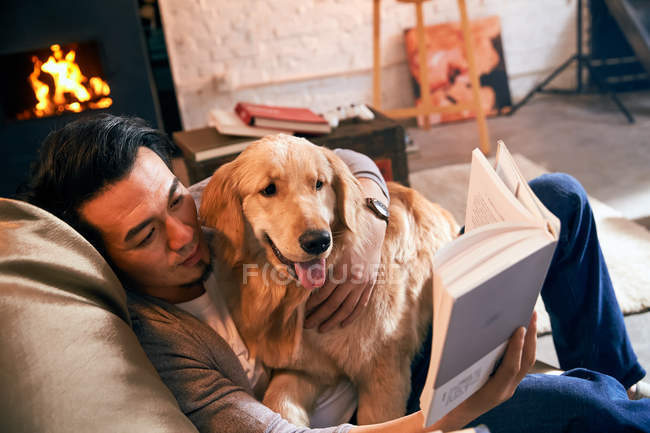 Schöner asiatischer Mann liest Buch, während er sich mit Hund zu Hause ausruht — Stockfoto