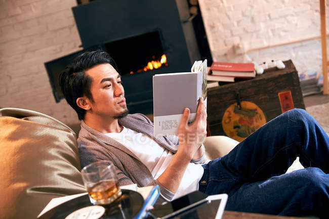 Concentré jeune asiatique homme reposant dans haricot sac chaise et lecture livre à la maison — Photo de stock