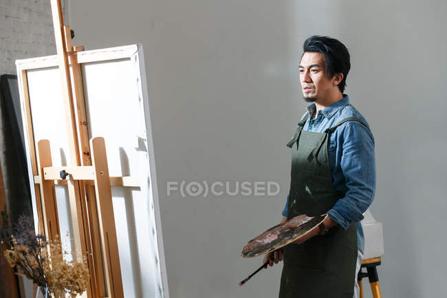 Концентрированный азиатский художник держит палитру и картину живописи в студии — стоковое фото