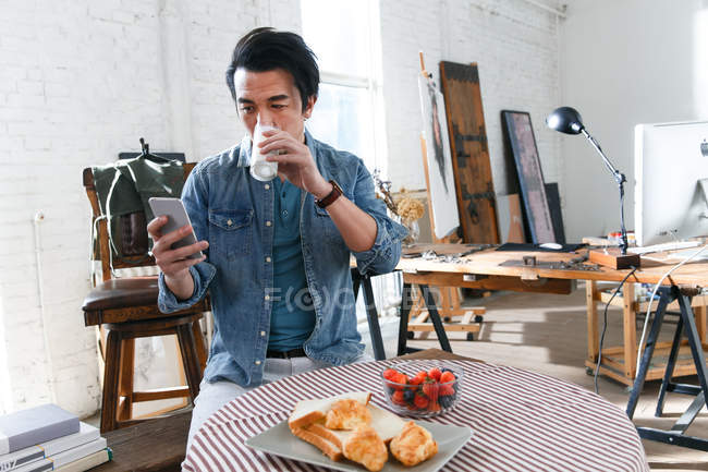 Junger asiatischer Mann trinkt Milch und benutzt Smartphone beim Frühstück im Kunstatelier — Stockfoto