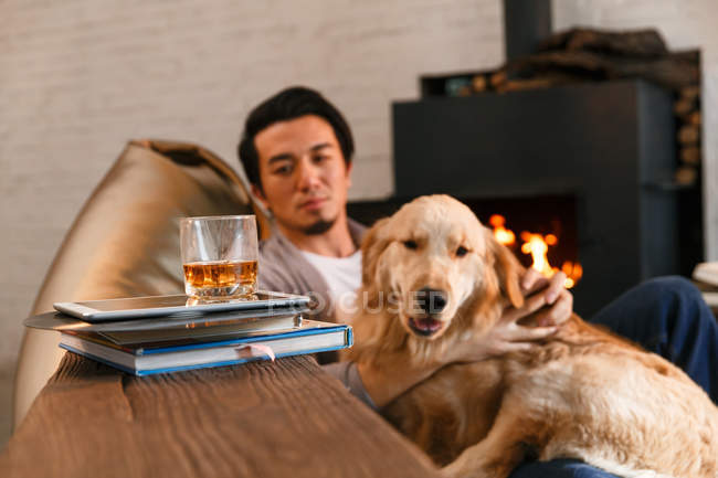 Glas Whisky mit digitalem Tablet und Büchern auf dem Tisch und asiatischer Mann ruht mit Hund zu Hause — Stockfoto