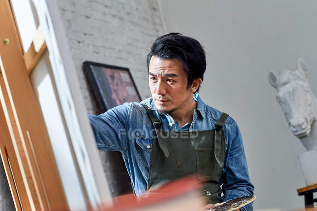 Selective focus of concentrated asian artist painting portrait in studio — Stock Photo
