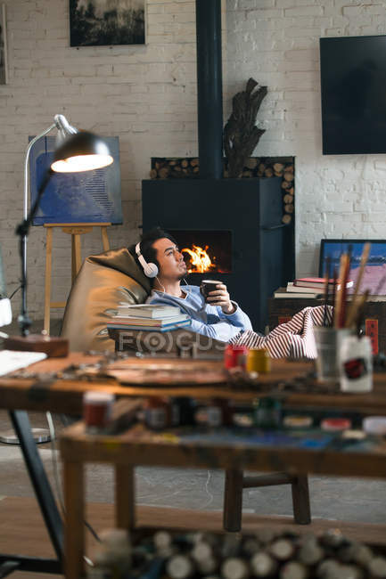 Vista de cerca de las herramientas de arte y hombre joven en los auriculares sentados cerca de la chimenea, enfoque selectivo - foto de stock