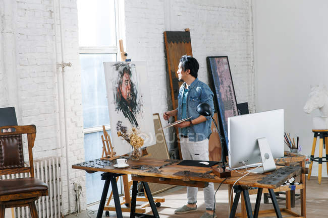 Фокусований чоловічий художник у фартусі, що тримає палітру та портрет живопису в студії — стокове фото