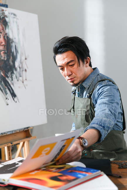 Ernsthafter asiatischer Maler in Schürze schaut sich Buch im Kunstatelier an — Stockfoto