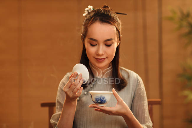 Bela sorrindo jovem chinesa segurando xícara e cheirando chá aromático — Fotografia de Stock