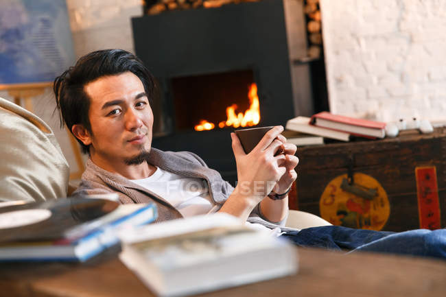 Schöner asiatischer Mann mit Tasse mit Heißgetränk und Blick auf Kamera zu Hause — Stockfoto