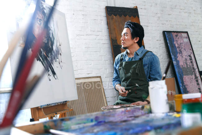 Selective focus of young male painter in apron looking at picture in studio — Stock Photo