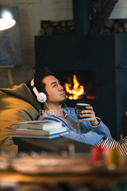 Задумчивый азиатский человек в наушниках держа чашку с горячим напитком и глядя в сторону дома — стоковое фото