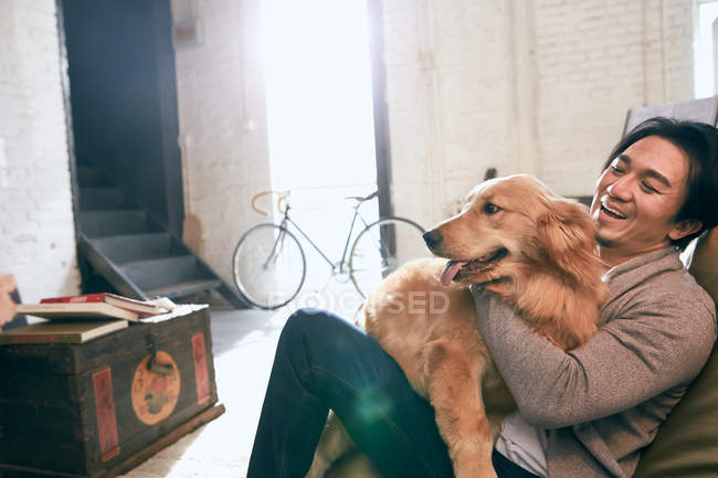 Handsome asian man sitting in bean bag chair and hugging dog at home — Stock Photo
