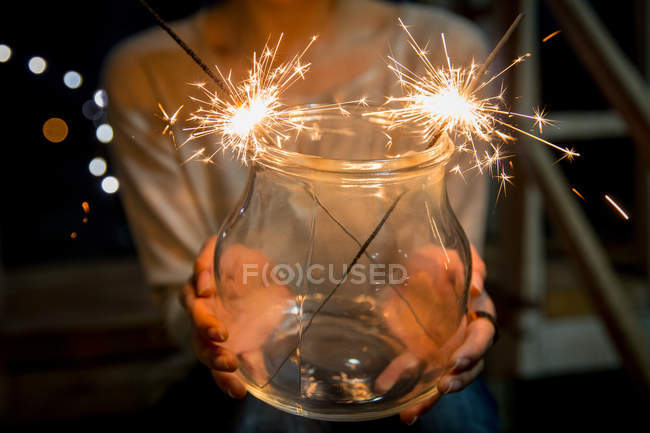 Tiro recortado de la persona sosteniendo frasco de vidrio con bengalas ardientes sobre fondo festivo borroso - foto de stock