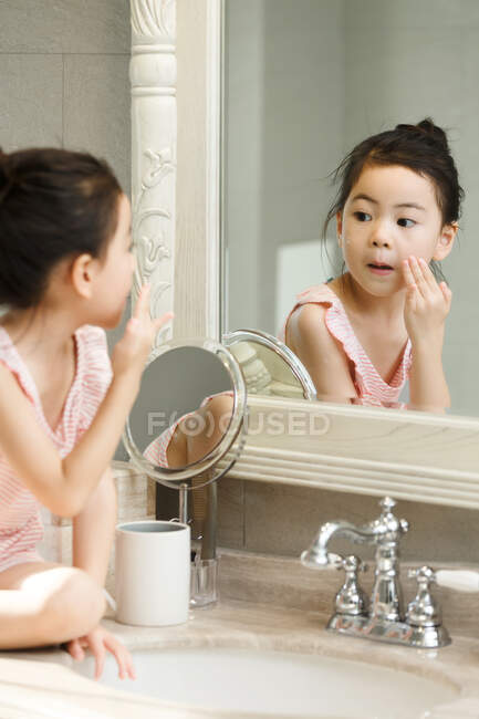 Little girl applying cream on her face in front of mirror — Stock Photo