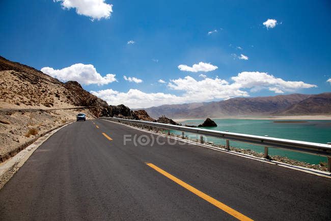 Coche conduciendo por carretera de asfalto cerca de la masa de agua y colinas escénicas en el Tíbet - foto de stock