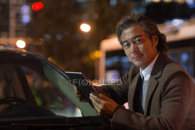 Mature asian man standing near car and using digital tablet in night city — Stock Photo