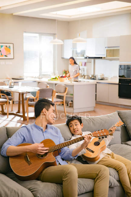 Vue grand angle du père et du fils heureux assis sur le canapé et jouant des guitares, la mère cuisinant derrière dans la cuisine — Photo de stock