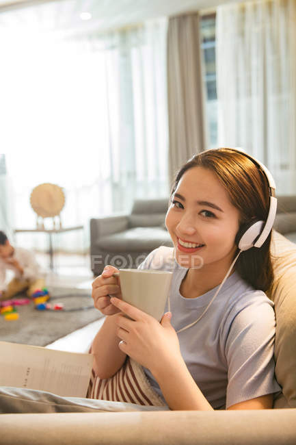 Chino mujer en auriculares celebración taza y sonriendo a la cámara mientras hijo jugando con juguetes detrás en casa - foto de stock