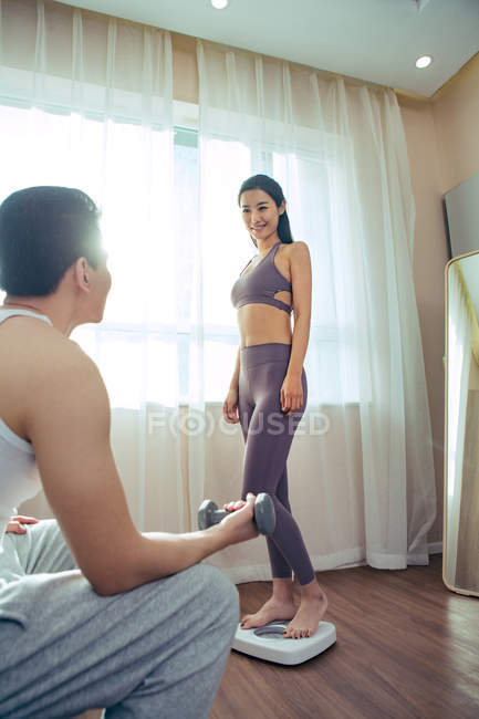 Улыбающаяся молодая женщина, стоящая на весах и глядя на бойфренда, упражняющегося с гантелями дома — стоковое фото