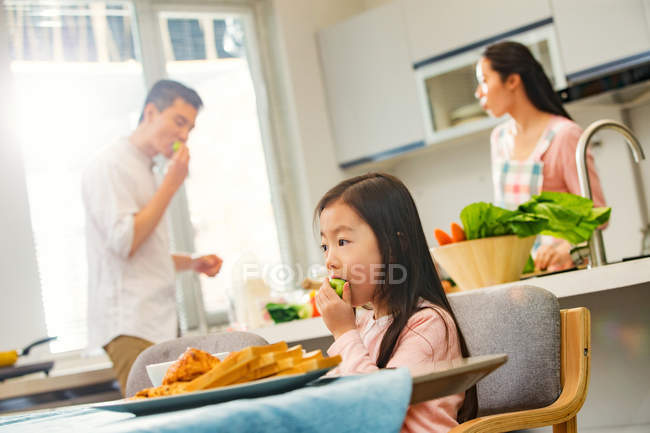 Adorabile bambino mangiare mela mentre i genitori in piedi dietro in cucina — Foto stock