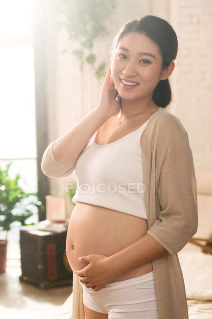 Allegra giovane donna incinta che tocca la pancia e sorride alla fotocamera a casa — Foto stock