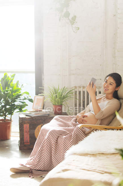 Lächelnde junge schwangere Frau sitzt im Schaukelstuhl und benutzt Smartphone — Stockfoto