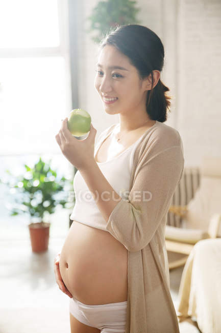 Улыбающаяся молодая беременная женщина держит зеленое яблоко и стоит дома — стоковое фото
