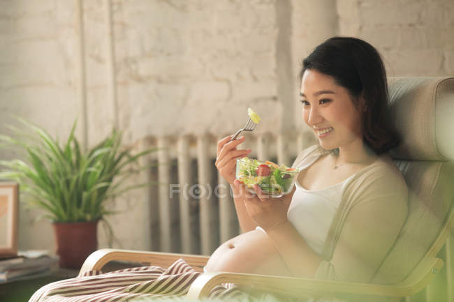 Улыбающаяся молодая беременная женщина сидит в кресле-качалке и ест овощной салат — стоковое фото