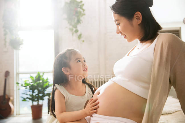 Feliz niña tocando el vientre de la sonriente madre embarazada en casa - foto de stock