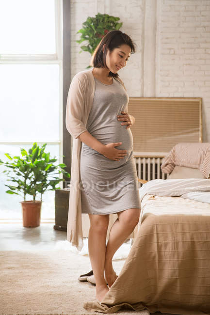 Schöne lächelnde junge schwangere Frau, die im Schlafzimmer steht und den Bauch berührt — Stockfoto