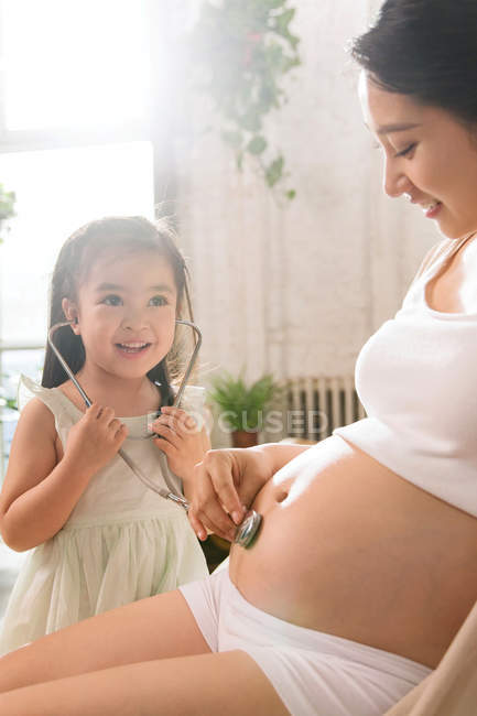 Adorável criança feliz segurando estetoscópio e brincando com a mãe grávida em casa — Fotografia de Stock