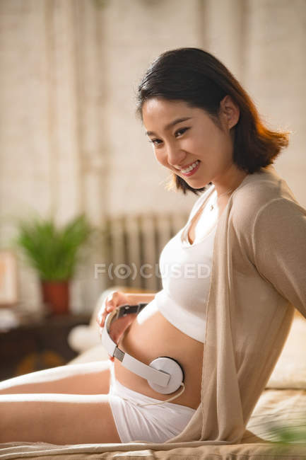 Vue latérale de la jeune femme enceinte souriante assise sur le lit et tenant des écouteurs sur le ventre — Photo de stock