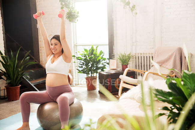Улыбающаяся молодая беременная женщина, сидящая на мяче для фитнеса и упражняющаяся с гантелями дома — стоковое фото