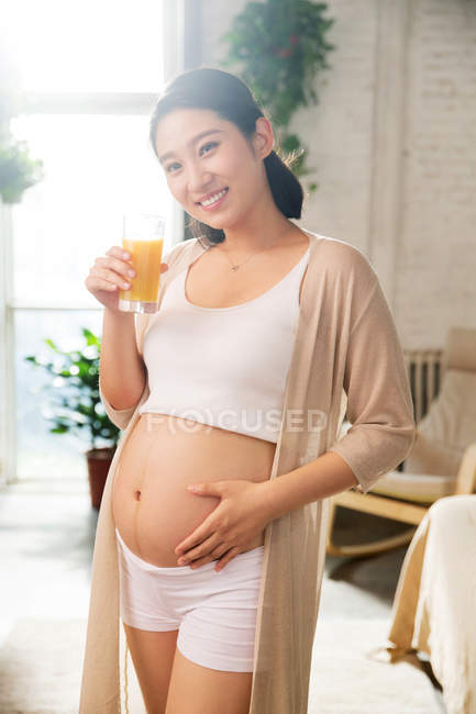 Feliz joven embarazada sosteniendo un vaso de jugo y sonriendo a la cámara - foto de stock