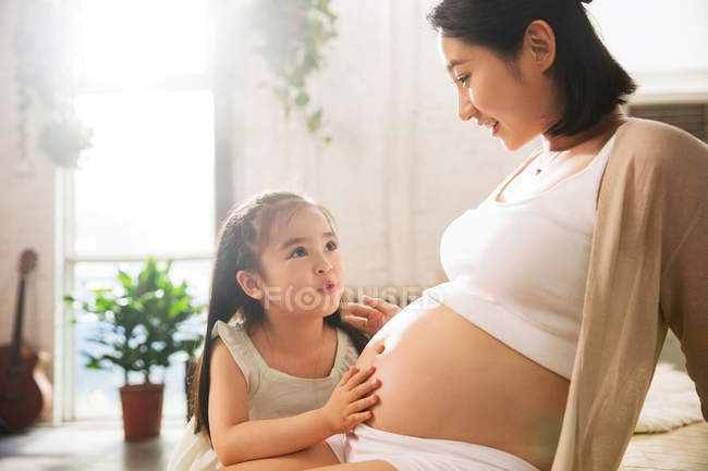 Feliz niña tocando el vientre de la sonriente madre embarazada en casa - foto de stock
