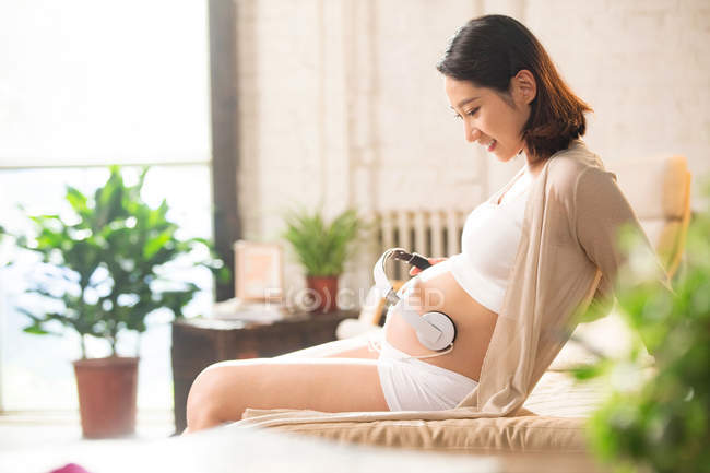 Вид сбоку улыбающейся молодой беременной женщины, сидящей на кровати и держащей наушники на животе — стоковое фото