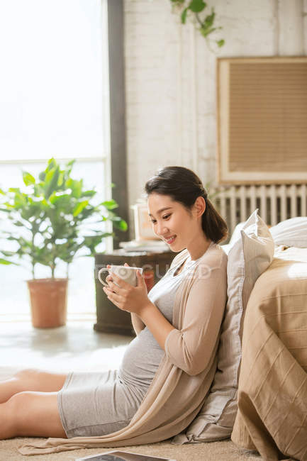 Вид збоку усміхненої молодої вагітної жінки, що тримає чашку з гарячим напоєм вдома — стокове фото