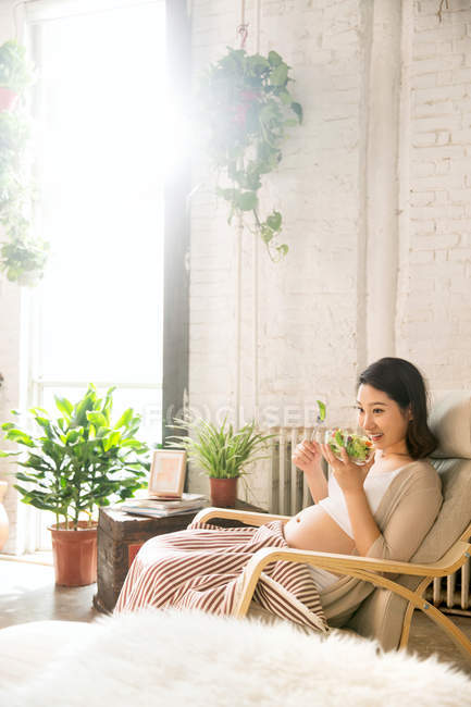Happy young pregnant woman sitting in rocking chair and holding bowl with vegetable salad — Stock Photo